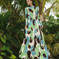 A woman stands outdoors before tropical plants, wearing the Cara Cara Aspen Dress—a longsleeve maxi featuring green, blue, white, and brown swirl patterns.