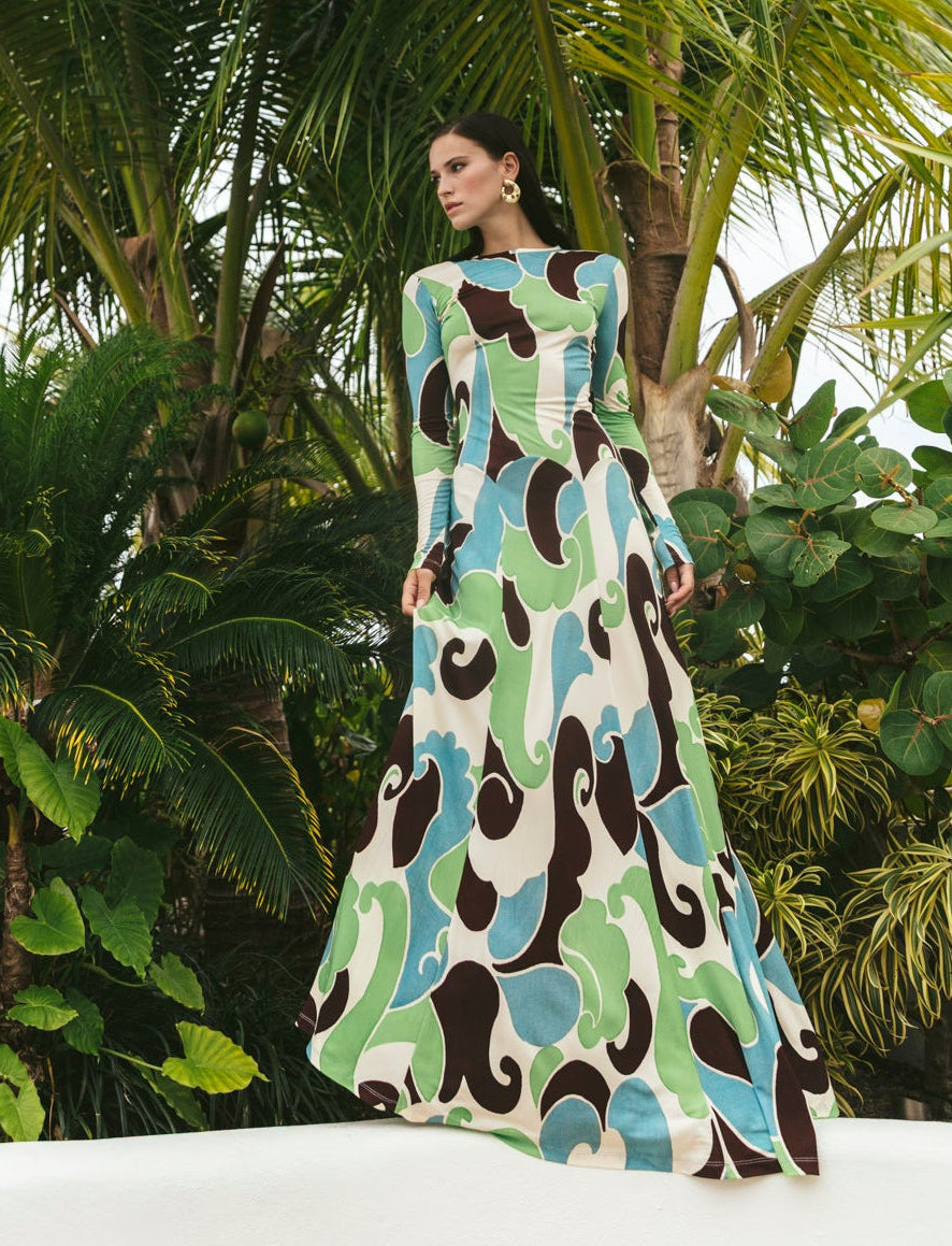 A woman stands outdoors before tropical plants, wearing the Cara Cara Aspen Dress—a longsleeve maxi featuring green, blue, white, and brown swirl patterns.