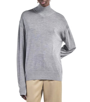 A person wearing a light heather grey Maria McManus Fine Gauge Mock Neck Sweater and beige pants stands with one hand on their hip against a white background.