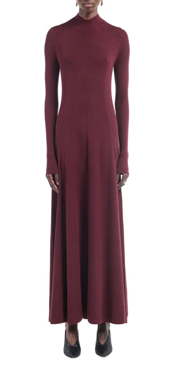 A person wearing the Maria McManus Rib Godet Dress—long-sleeved, ankle-length merlot with a fitted bodice and high neckline—and black shoes, stands against a white background.