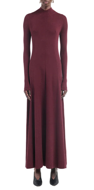 A person wearing the Maria McManus Rib Godet Dress—long-sleeved, ankle-length merlot with a fitted bodice and high neckline—and black shoes, stands against a white background.