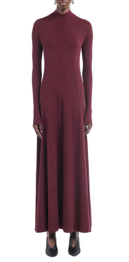 A person wearing the Maria McManus Rib Godet Dress—long-sleeved, ankle-length merlot with a fitted bodice and high neckline—and black shoes, stands against a white background.