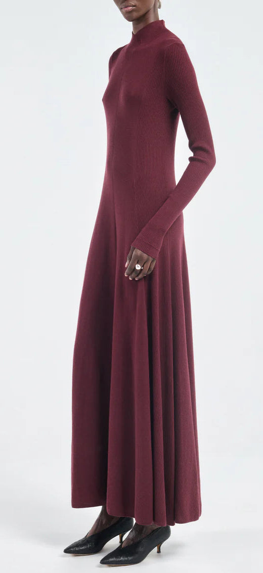 A person in the Maria McManus Rib Godet Dress—long-sleeved, floor-length merlot—with black pointed shoes stands against a plain light background.