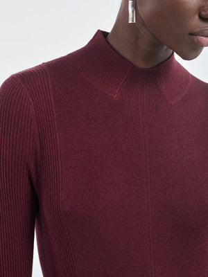 A person wearing the Maria McManus Rib Godet Dress in burgundy merlot with long sleeves, a mock neckline, and geometric silver earrings.
