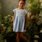 A young girl wearing the Louise Misha Girls’ Briska Dress in light blue and yellow sandals stands among tall wildflowers, set against a neutral gray wall.