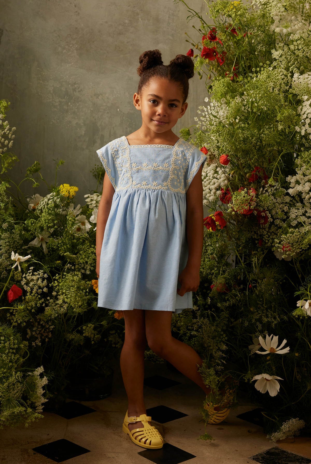 A young girl wearing the Louise Misha Girls’ Briska Dress in light blue and yellow sandals stands among tall wildflowers, set against a neutral gray wall.