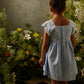A young girl, dressed in the Louise Misha Girls’ Briska Dress, stands indoors on a tiled floor with her back to the camera, gazing at a display of wildflowers set against a textured wall.