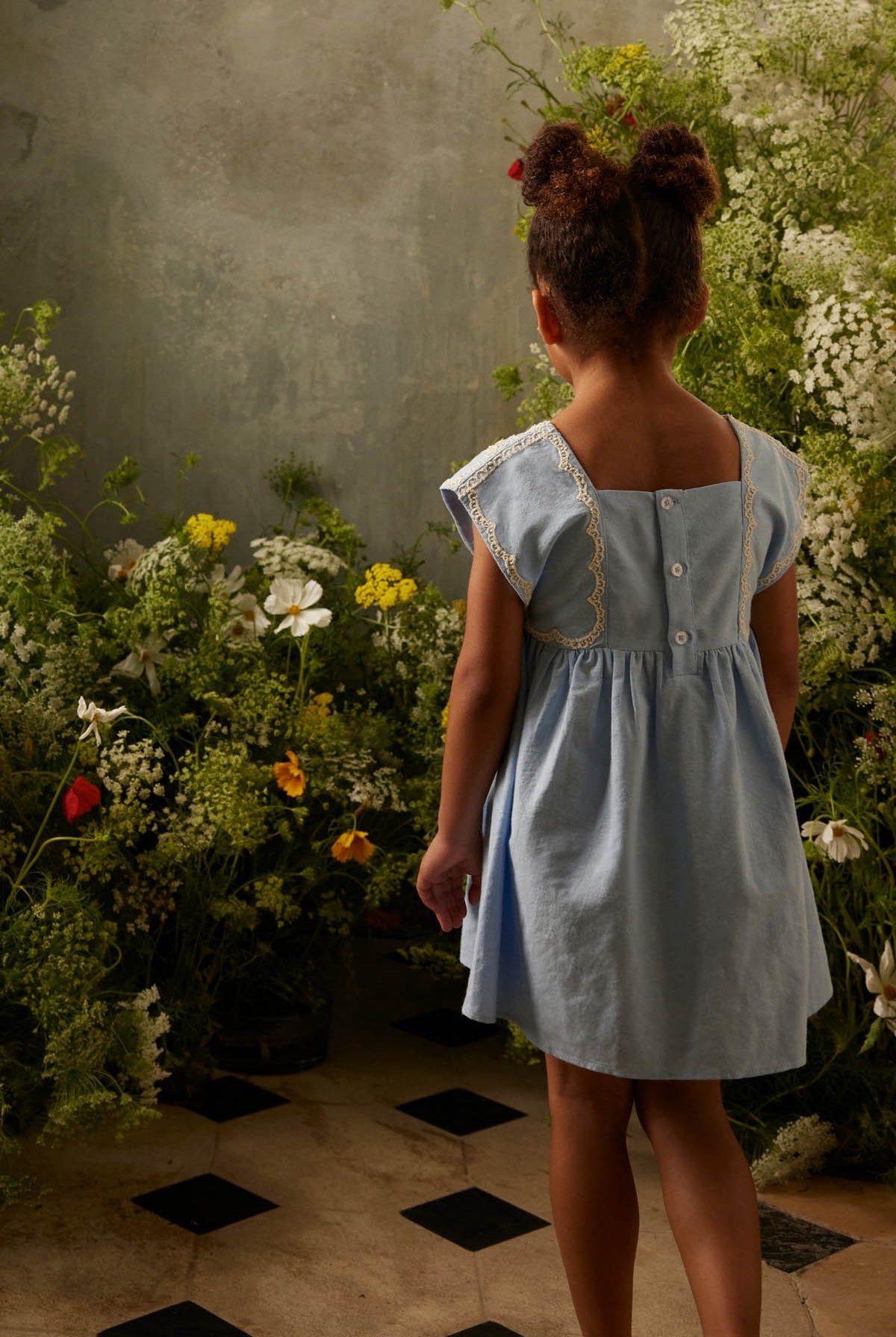 A young girl, dressed in the Louise Misha Girls’ Briska Dress, stands indoors on a tiled floor with her back to the camera, gazing at a display of wildflowers set against a textured wall.