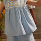 A young child is wearing Louise Misha’s Girls’ Vally Shorts, crafted from Oxford cotton with an elasticized waist, paired with a light blue embroidered sleeveless top featuring delicate detailing, while standing indoors with another child partially visible in the background.