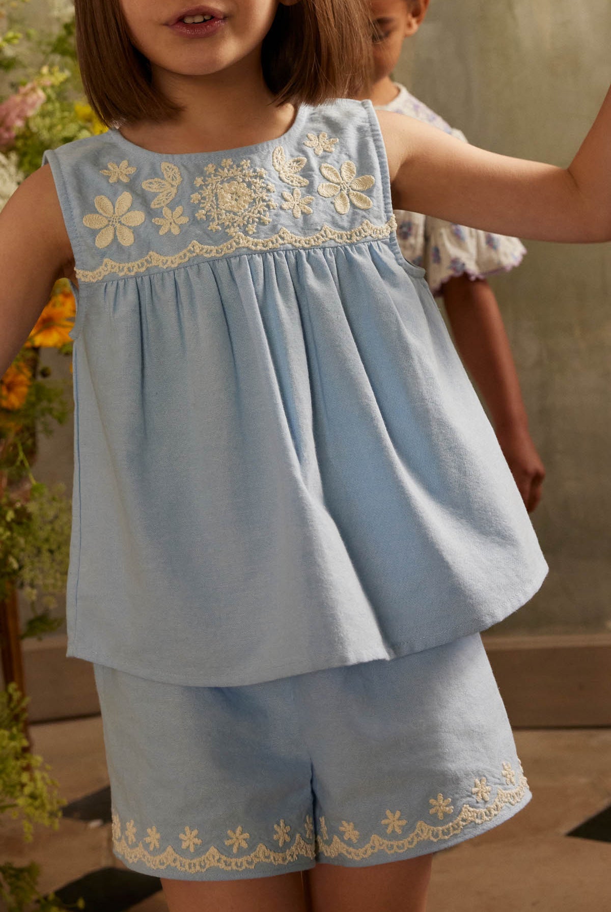 A young child is wearing Louise Misha’s Girls’ Vally Shorts, crafted from Oxford cotton with an elasticized waist, paired with a light blue embroidered sleeveless top featuring delicate detailing, while standing indoors with another child partially visible in the background.