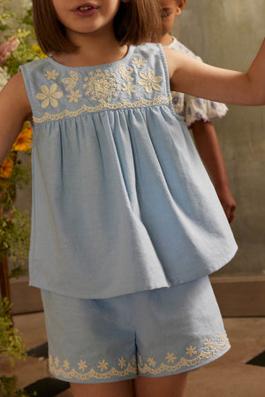 A young child is wearing Louise Misha’s Girls’ Vally Shorts, crafted from Oxford cotton with an elasticized waist, paired with a light blue embroidered sleeveless top featuring delicate detailing, while standing indoors with another child partially visible in the background.
