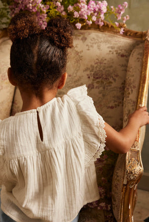 A child with two buns, wearing the Louise Misha Girls’ Maiya Blouse in white cotton gauze, sits on a floral upholstered chair facing away. Pink flowers are visible in the background.