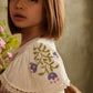 A young girl with straight brown hair stands indoors near a bouquet of flowers, wearing the Louise Misha Girls’ Ana Dress—a white cotton dress adorned with embroidered flowers and delicate lace trim.