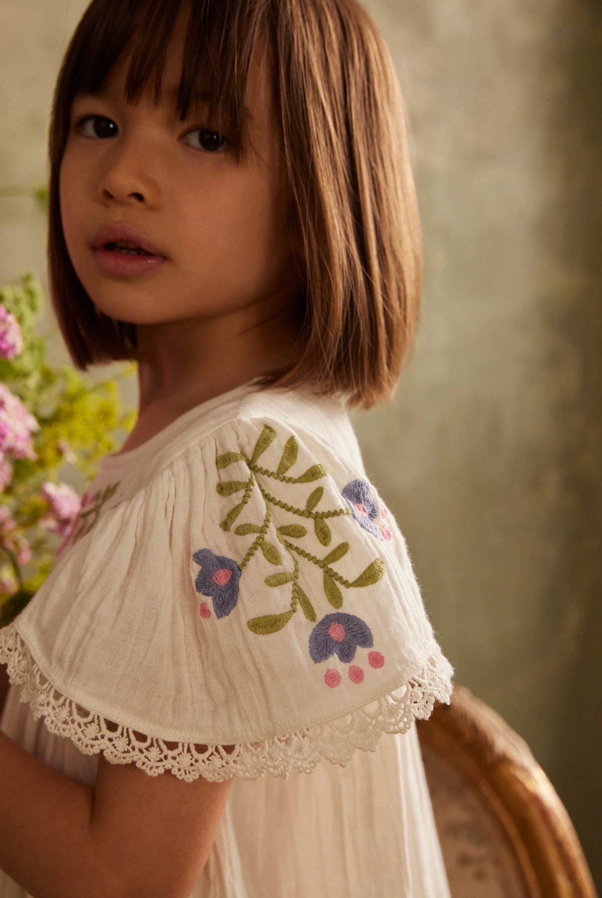 A young girl with straight brown hair stands indoors near a bouquet of flowers, wearing the Louise Misha Girls’ Ana Dress—a white cotton dress adorned with embroidered flowers and delicate lace trim.