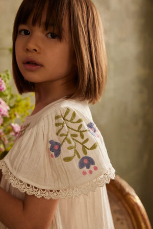 A young girl with straight brown hair stands indoors near a bouquet of flowers, wearing the Louise Misha Girls’ Ana Dress—a white cotton dress adorned with embroidered flowers and delicate lace trim.