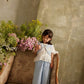 A young girl models the Louise Misha Girls’ Florelia Pants, featuring delicate embroidered details, as she stands on a vintage floral sofa surrounded by green and purple flowers against a textured wall.