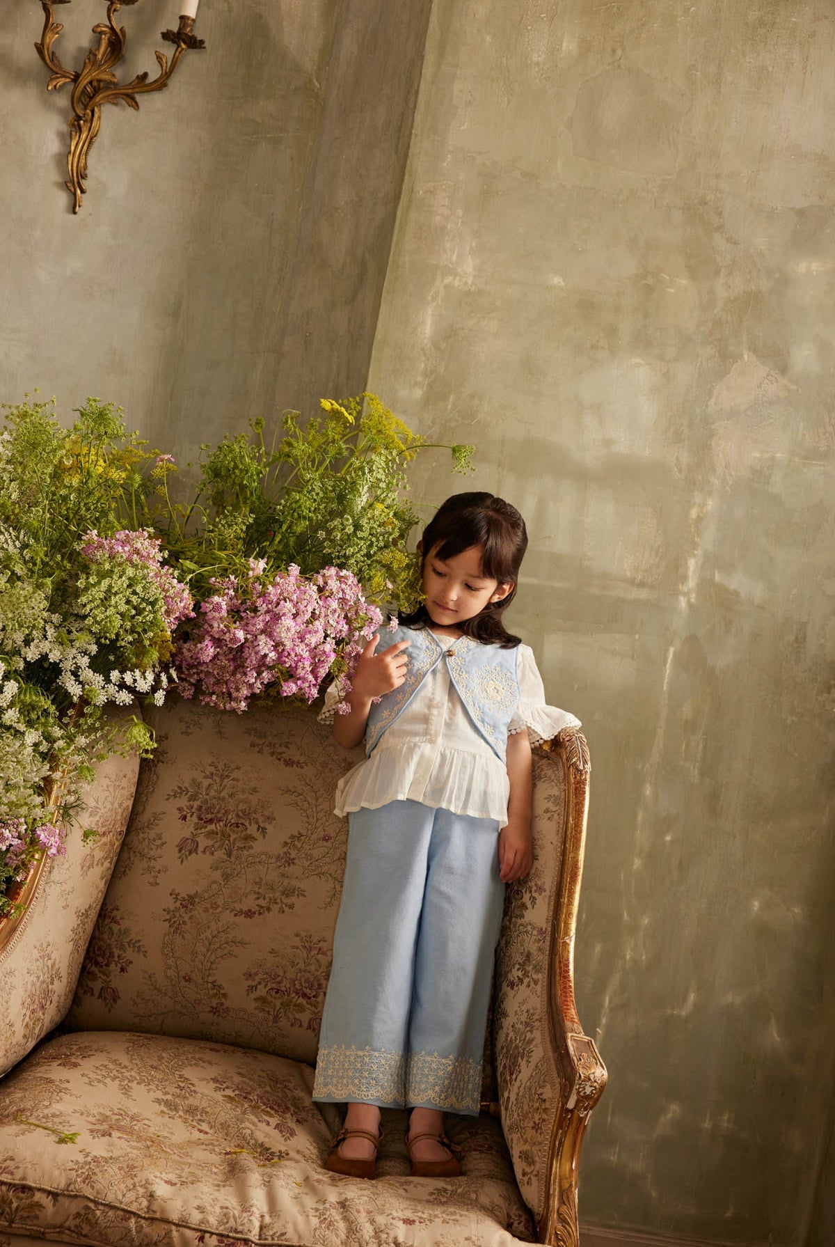 A young girl models the Louise Misha Girls’ Florelia Pants, featuring delicate embroidered details, as she stands on a vintage floral sofa surrounded by green and purple flowers against a textured wall.