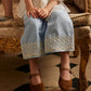A child sits on an ornate sofa, wearing Louise Misha’s Girls’ Florelia Pants—light blue Oxford cotton trousers with an elasticized waist and delicate embroidered details—paired with brown Mary Jane shoes, hands folded neatly on their lap.