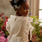 A young girl with flowers in her hair wears the Louise Misha Girls’ Esia Blouse, a white eyelet embroidered top, while standing among colorful flowers as soft sunlight streams through a nearby window.