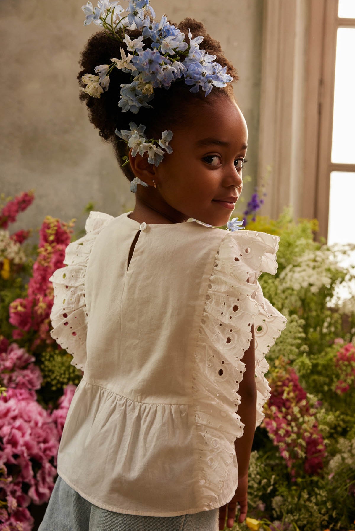 A young girl with flowers in her hair wears the Louise Misha Girls’ Esia Blouse, a white eyelet embroidered top, while standing among colorful flowers as soft sunlight streams through a nearby window.