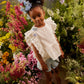 A young girl stands among colorful flowers, wearing the Louise Misha Girls’ Esia Blouse, denim shorts, brown sandals, and blue flowers in her hair, looking at the camera.