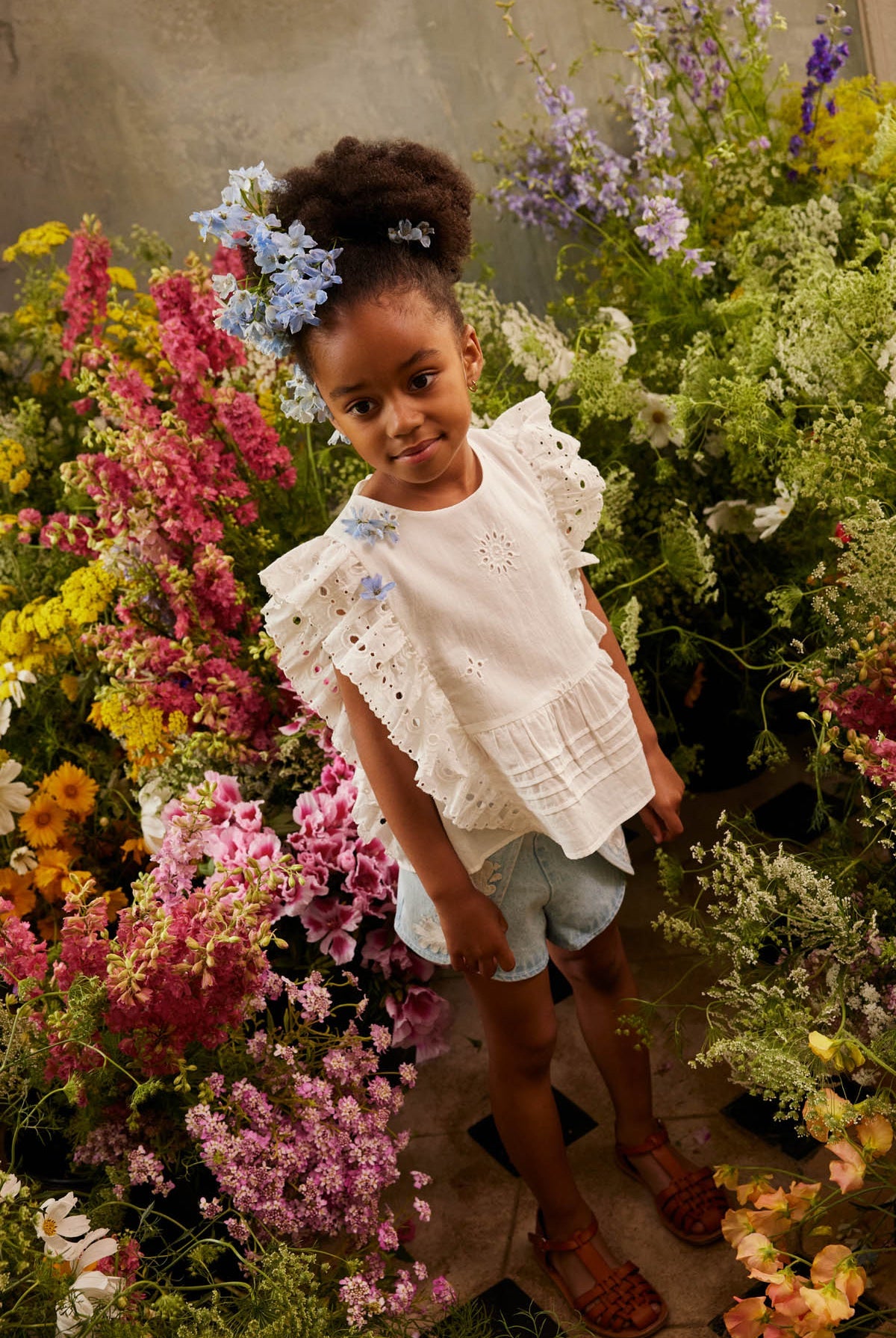 A young girl stands among colorful flowers, wearing the Louise Misha Girls’ Esia Blouse, denim shorts, brown sandals, and blue flowers in her hair, looking at the camera.