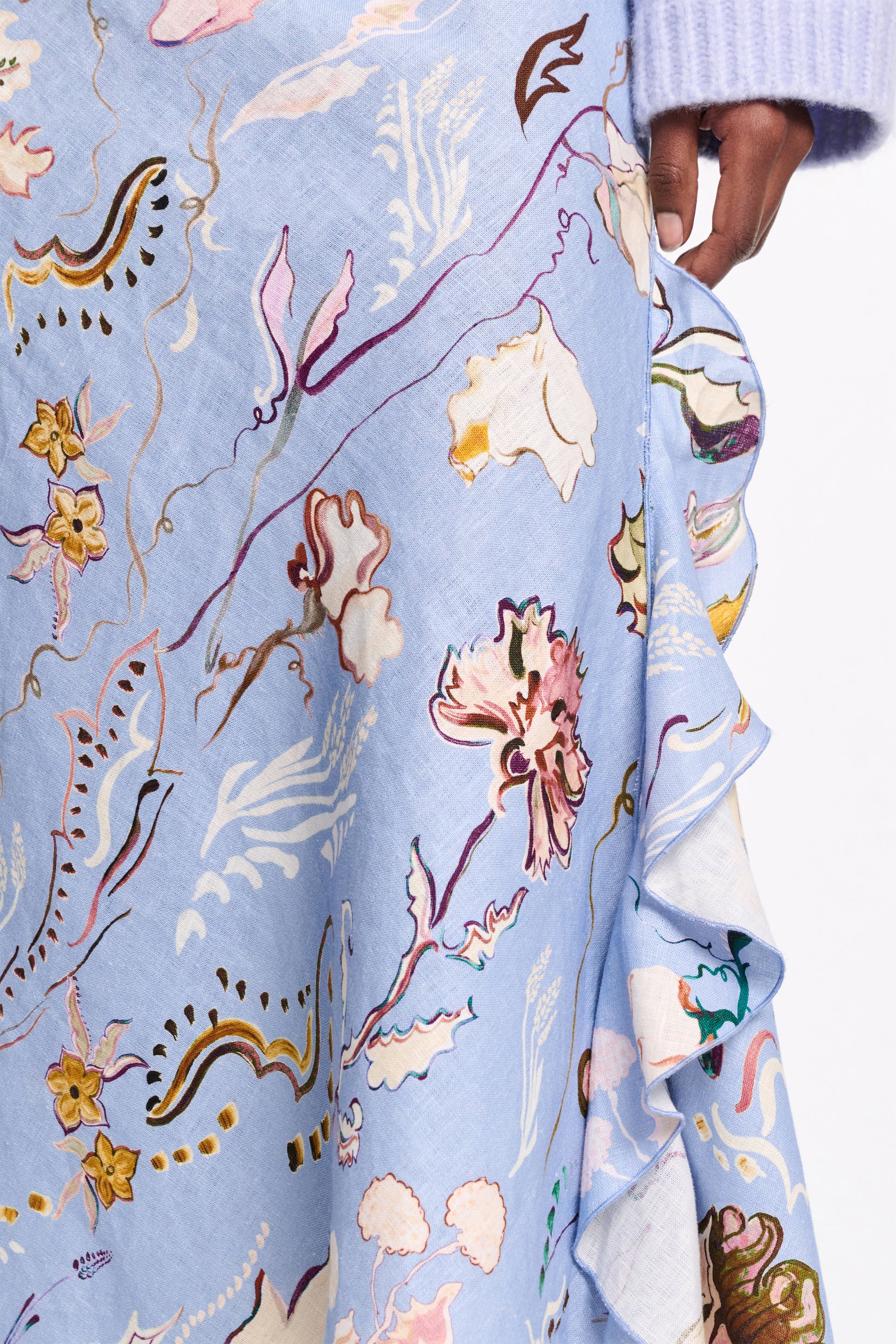 A close-up of a person wearing the Dorothee Schumacher Floral Power Skirt in light blue linen, featuring a vibrant floral pattern and an asymmetric cut. The look is finished with a light purple sleeve.