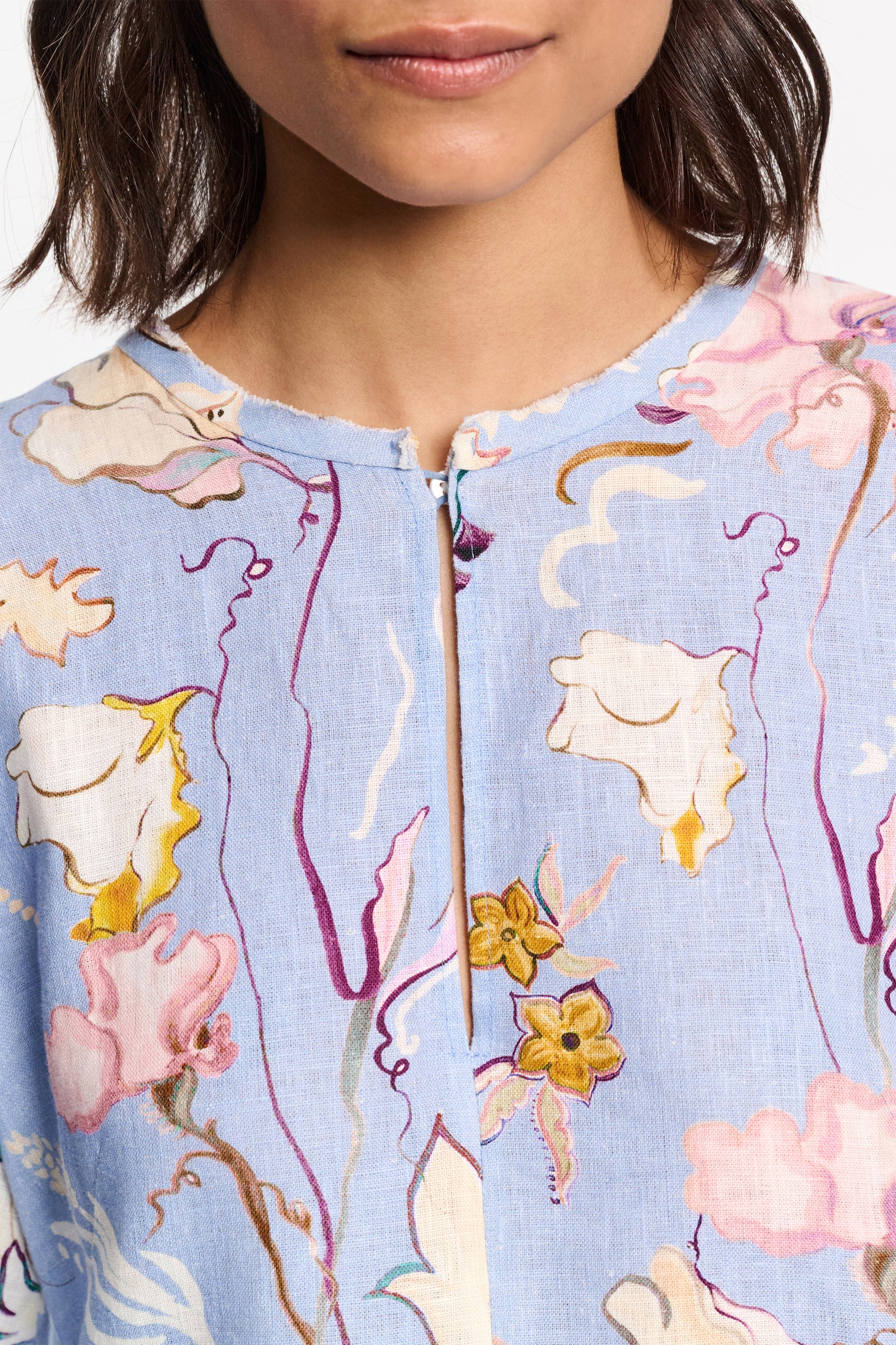 Close-up of someone in a Dorothee Schumacher Floral Power Mini Dress. The blue linen shift dress features a round neckline with pink, yellow, and white floral patterns. Voluminous sleeves add elegance, while the person's head is partially visible.