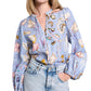 A person is styled in the Dorothee Schumacher Floral Power Blouse, featuring a V-neckline and long sleeves. They pair it with light blue jeans and a silk georgette scarf, effortlessly draping around their neck for an elegant touch, while posing with one hand in the pocket.