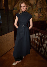 A woman stands indoors on a wooden floor, hands in her pockets, wearing the Adam Lippes Akane Dress over black pants with white shoes.