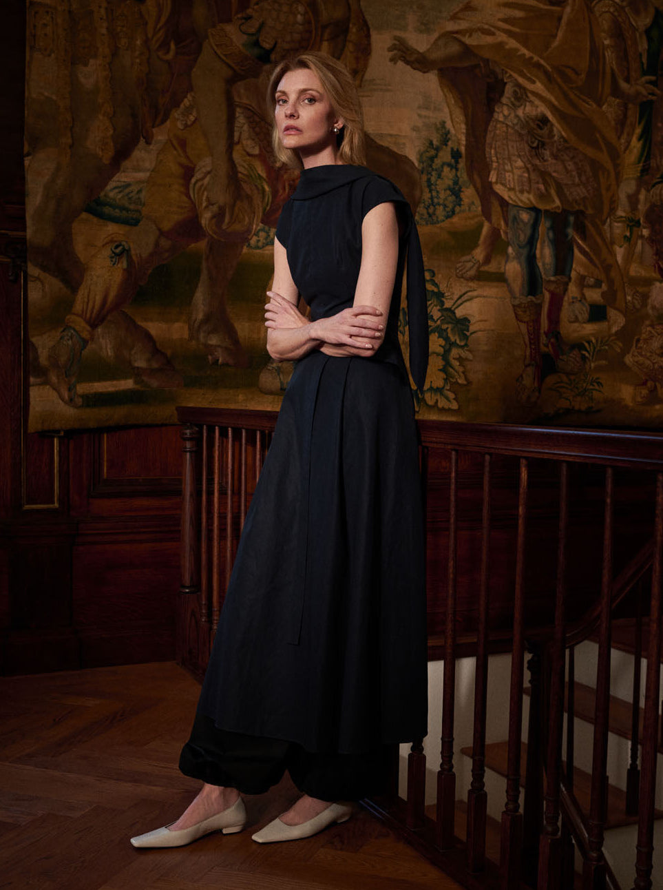 A woman in the sleeveless Akane Dress by Adam Lippes, featuring an A-line skirt, stands by a wooden railing with a tapestry of figures in the background and wears white shoes.
