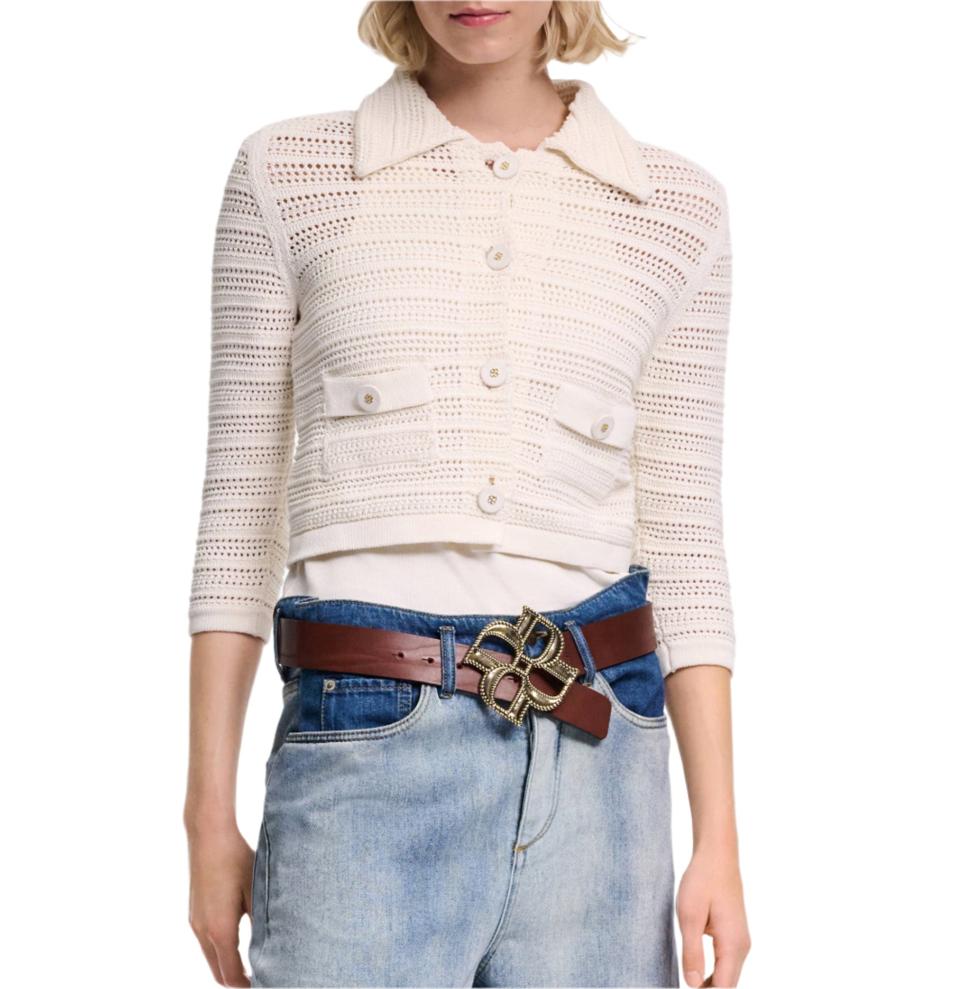 A woman wears the Dorothee Schumacher Precise Pointelle Jacket—a white crocheted button-up cardigan with graphic pointelle details—paired with a white top, blue jeans, and a brown belt featuring a large decorative buckle.