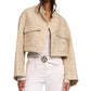A woman wears the Dorothee Schumacher Shiny Coolness Jacket, a beige Lurex-tweed piece with large front pockets, styled with a white shirt, pants, and a gold-buckled belt for a chic boxy look.