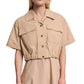 A woman wears the Poplin Power Cropped Blouse by Dorothee Schumacher, featuring beige cotton poplin, short sleeves, chest pockets, an elasticated waist, and an open collar.