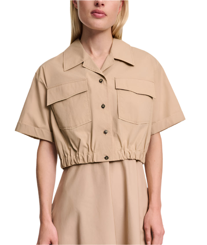 A woman wears the Poplin Power Cropped Blouse by Dorothee Schumacher, featuring beige cotton poplin, short sleeves, chest pockets, an elasticated waist, and an open collar.