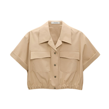 The Poplin Power Cropped Blouse by Dorothee Schumacher is a beige cotton poplin top featuring short sleeves, an open collar, two large chest pockets, and an elastic hem for a cropped fit.