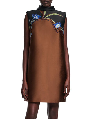 A person stands with arms at their sides, wearing Dorothee Schumacher’s Soft Petal Dress—a sleeveless brown shift featuring a black stand collar and blue-green floral accents near the shoulders, highlighting Italian artistry.