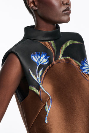 A person wearing the Soft Petal Dress by Dorothee Schumacher, a sleeveless brown and black stand collar cloque jacquard dress with blue and white floral embroidery on the chest that highlights Italian artistry.