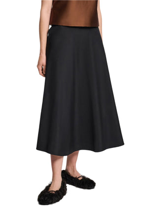 A person wearing a brown sleeveless top, Dorothee Schumacher's Sporty Elegance Skirt in black, and black fuzzy slip-on shoes stands against a white background.