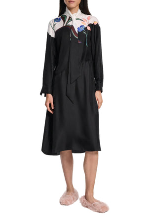A woman wears the Seductive Bloom Maxi Dress by Dorothee Schumacher, a black dress with floral print on the shoulder and chest and a wrap-around tie collar, paired with pink fuzzy slippers.
