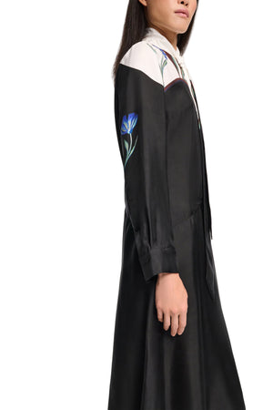 A woman wears the Seductive Bloom Maxi Dress by Dorothee Schumacher—a long-sleeved black silk shirtdress with a white yoke and blue floral embroidery—standing against a plain light background.