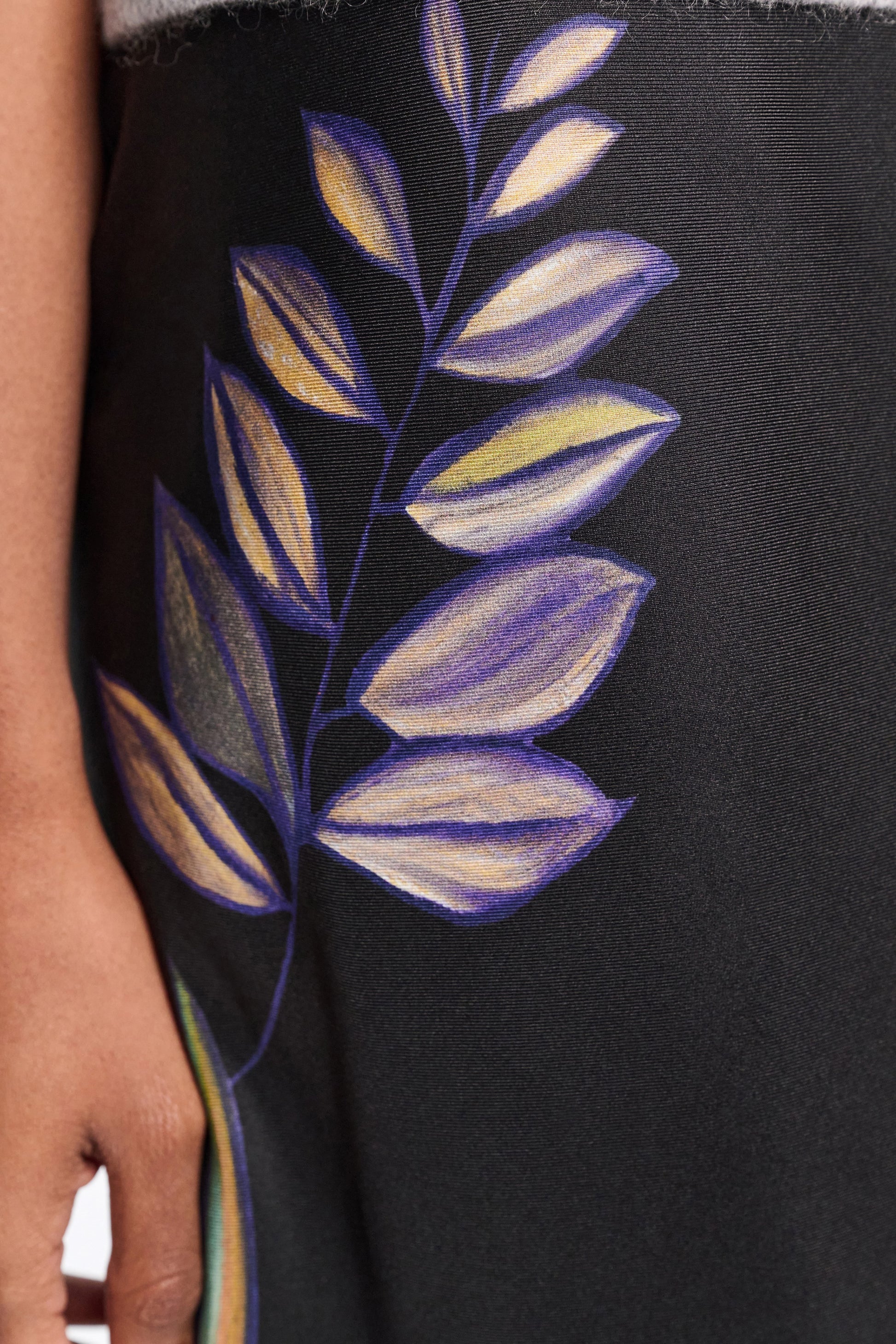 A close-up of a hand beside the Dorothee Schumacher Seductive Bloom Skirt, a black silk midi-skirt with a painted purple and yellow leaf design.