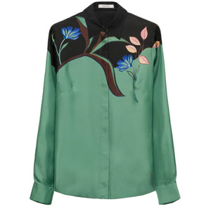 The Seductive Bloom Blouse by Dorothee Schumacher is a silk twill, long-sleeve, button-up top featuring a green body, black shoulders, and a floral print with blue flowers and pink leaves on the upper front.