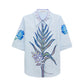 The Floral Embrace Blouse by Dorothee Schumacher is crafted in light blue cotton-poplin, featuring botanical prints on the front, floral designs on the sleeves, and leaf patterns on the collar.