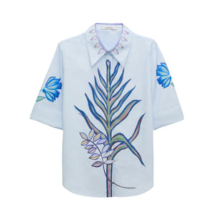 The Floral Embrace Blouse by Dorothee Schumacher is crafted in light blue cotton-poplin, featuring botanical prints on the front, floral designs on the sleeves, and leaf patterns on the collar.