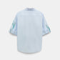 The Floral Embrace Blouse by Dorothee Schumacher is a light blue short-sleeve classic shirt, shown from the back, with subtle green and purple floral designs on the sleeves and collar—a fresh twist on a timeless print.