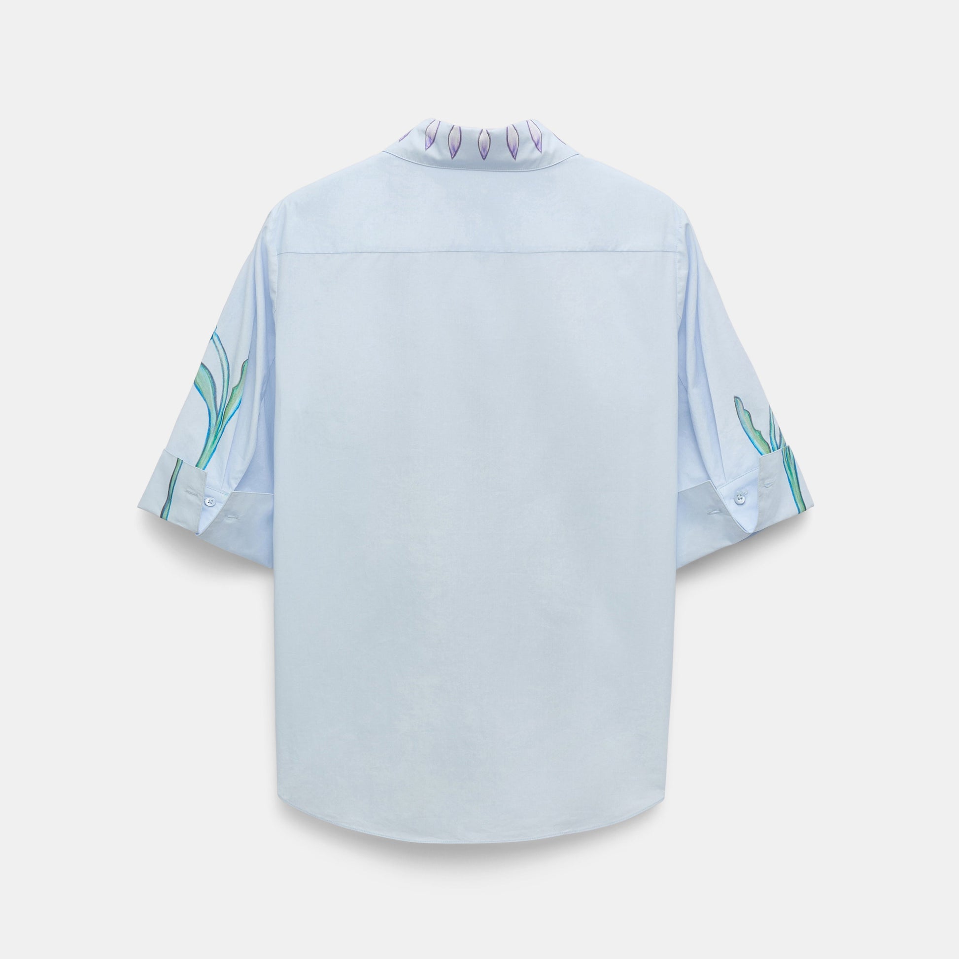 The Floral Embrace Blouse by Dorothee Schumacher is a light blue short-sleeve classic shirt, shown from the back, with subtle green and purple floral designs on the sleeves and collar—a fresh twist on a timeless print.