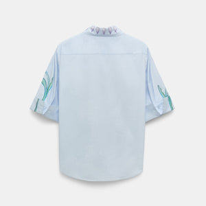 The Floral Embrace Blouse by Dorothee Schumacher is a light blue short-sleeve classic shirt, shown from the back, with subtle green and purple floral designs on the sleeves and collar—a fresh twist on a timeless print.