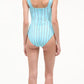 A woman with long blonde hair, wearing the Staud Adelaide One Piece—a light blue swimsuit with white vertical stripes, supportive straps, and back buttons—stands in flip-flops facing away.