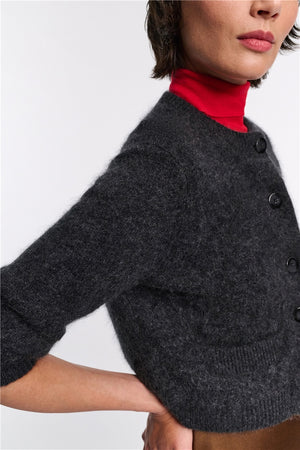 Someone wears the Dorothee Schumacher Fluffy Statements Cardigan in dark gray over a red turtleneck sweater, with their arm bent and hand resting on their hip.